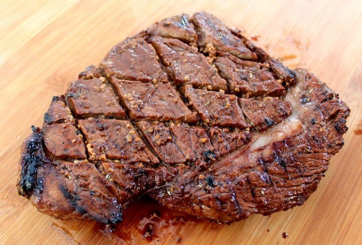 A marinated steak rests on a wooden cutting board after being cooked on a grill