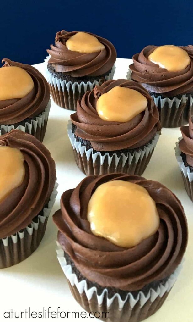 An image of chocolate cupcakes with chocolate fudge frosting and a salted caramel center on a white serving tray  against a blue wall. These Chocolate Salted Caramel Cupcakes are easy to make but taste decadent!