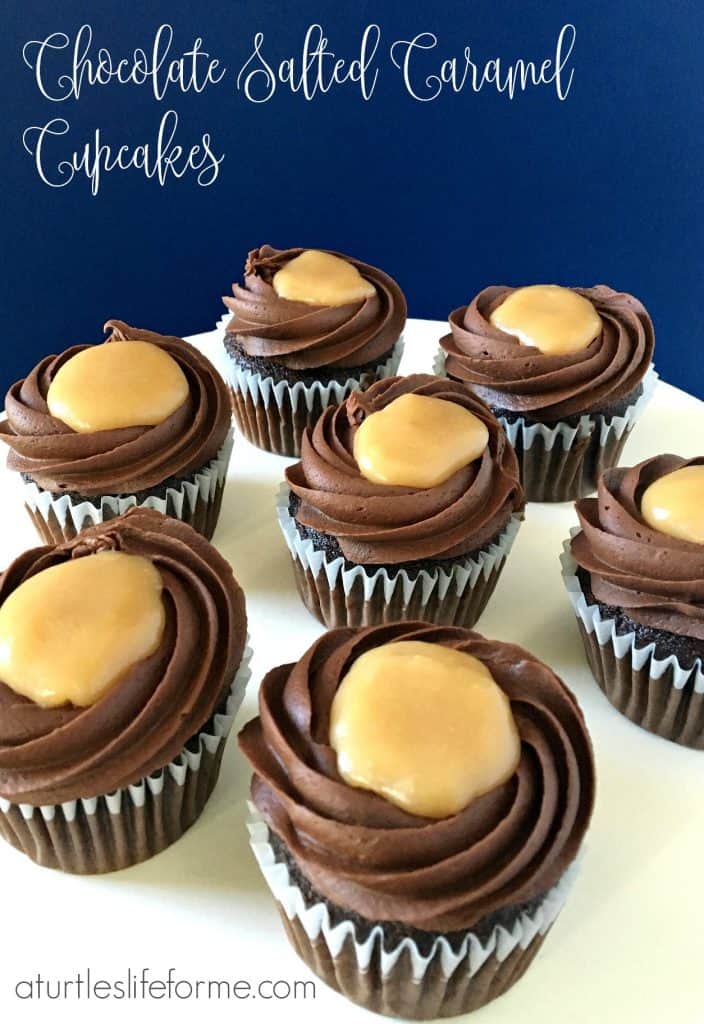 An image of chocolate cupcakes with chocolate fudge frosting and a salted caramel center on a white serving tray  against a blue wall. These Chocolate Salted Caramel Cupcakes are easy to make but taste decadent!